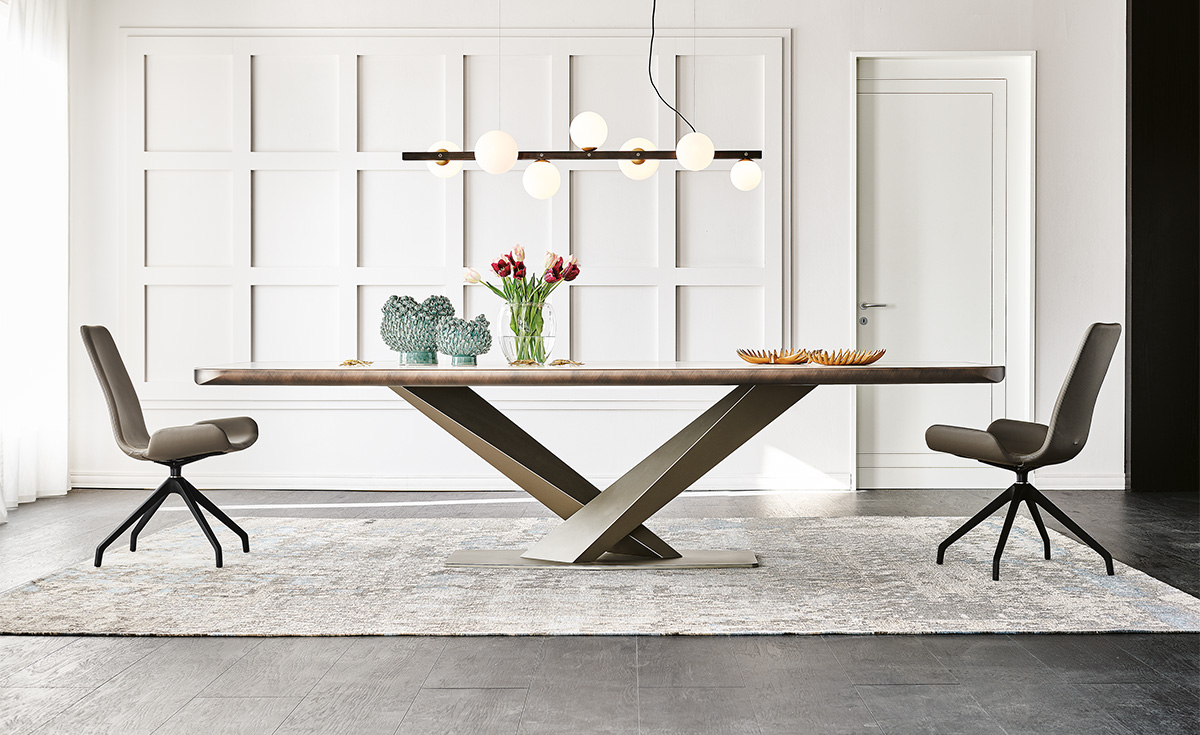 Elegantní jídelna, která oslavuje minimalismus a moderní design. Stůl Stratos s křížovým podstavcem a otočné jídelní židle s jemnými liniemi vytvářejí čistý, ale výrazný vizuální dojem, zvýrazněný geometrickým svítidlem a teplými dekoracemi.